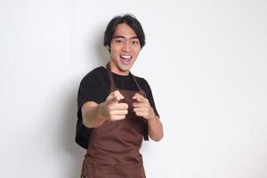 Portrait of attractive Asian barista man in brown apron showing product, pointing at something with hands. Advertising concept. Isolated image on white background photo