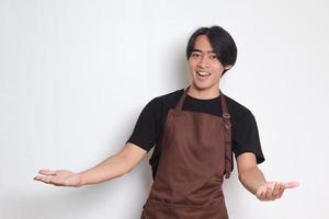 Portrait of attractive Asian barista man in brown apron spreading arms, inviting customer to come in. Advertising concept. Isolated image on white background photo