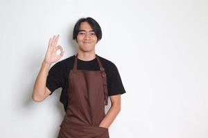 retrato de atractivo asiático barista hombre en marrón delantal demostración Okay mano gesto y sonriente mirando a cámara. publicidad concepto. aislado imagen en blanco antecedentes foto