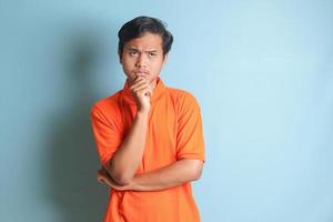 confused Asian man in orange shirt standing against blue background, thinking about question with hand on chin photo