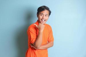 attractive Asian man in orange shirt with thumb pointing away on empty space. Advertising concept. Isolated image on blue background photo