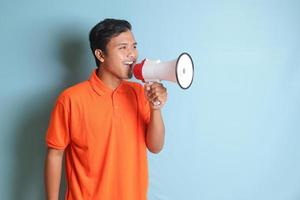 retrato de atractivo asiático hombre en naranja camisa Hablando mas fuerte utilizando megáfono, promoviendo producto. publicidad concepto. aislado imagen en azul antecedentes foto
