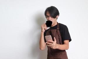 Portrait of attractive Asian barista man in brown apron drinking coffee while using mobile phone. Isolated image on white background photo