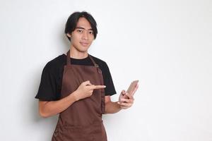 Portrait of attractive Asian barista man in brown apron holding and pointing mobile phone. Isolated image on white background photo