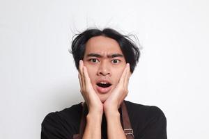 Close up portrait of shocked Asian barista man in brown apron holding hands on cheeks, feeling surprised with open mouth. Isolated image on white background photo