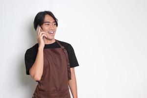 Portrait of attractive Asian barista man in brown apron having conversation while calling someone on mobile phone. Isolated image on white background photo