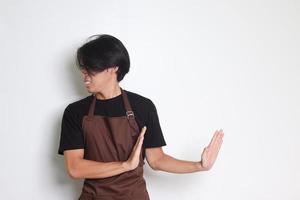 Portrait of attractive Asian barista man in brown apron forming a hand gesture to avoid something. Isolated image on white background photo