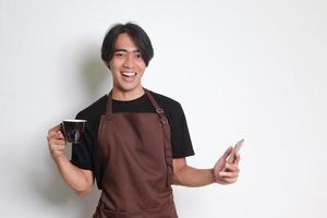 retrato de atractivo asiático barista hombre en marrón delantal participación un taza de café mientras utilizando móvil teléfono y sonriente a cámara. aislado imagen en blanco antecedentes foto