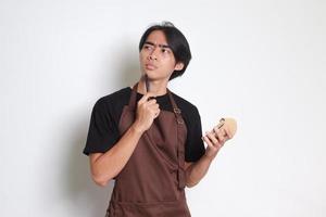 Portrait of attractive Asian barista man in brown apron taking order, writing on menu book list, thinking with pen on chin. Isolated image on white background photo