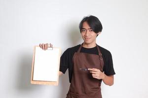 Portrait of attractive Asian barista man in brown apron showing blank paper of menu on clipboard. Isolated image on white background photo