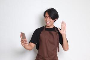 Portrait of attractive Asian barista man in brown apron making hi gesture while having conversation on video call. Isolated image on white background photo