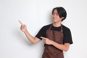 Portrait of attractive Asian barista man in brown apron showing product, pointing at something with hands. Advertising concept. Isolated image on white background photo