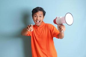 Portrait of attractive Asian man in orange shirt speaking louder using megaphone, promoting product. Advertising concept. Isolated image on blue background photo