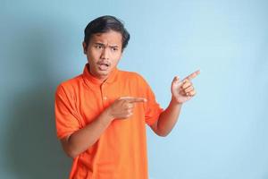 excited Asian man in orange shirt smiling and looking at the camera pointing with two hands and fingers to the side. Isolated image on blue background photo