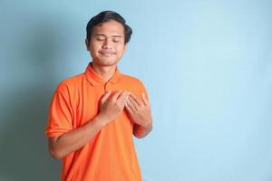 calming Asian man in orange shirt placing hand on chest and feeling peaceful. Mental health day concept, expresses sympathy and love, smiles positively. Isolated image on blue background photo