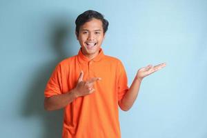 excited Asian man in orange shirt smiling and looking at the camera pointing with two hands and fingers to the side. Isolated image on blue background photo