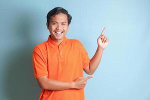 excited Asian man in orange shirt smiling and looking at the camera pointing with two hands and fingers to the side. Isolated image on blue background photo