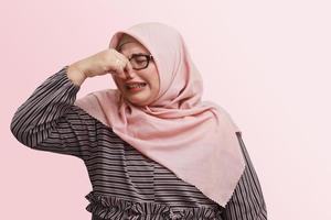 Portrait of unpleasant Asian woman with hijab, pinches nose with fingers hands looks with disgust something stinks bad smell situation. Isolated image on pink background photo