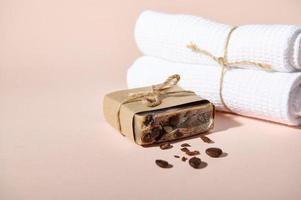 Organic homemade bar of soap with ground and scattered coffee beans and rolled towels, isolated on pink background photo