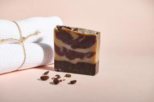 A bar of natural organic soap or solid body scrub, with ground coffee beans next to a white bath towel on pink backdrop photo