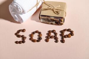 Top view of coffee beans laid out in SOAP word on pink background with natural soap bar - a body scrub and white towel. photo