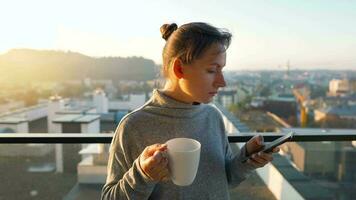 mujer empieza su día con un taza de té o café y comprobación correos electrónicos en su teléfono inteligente en el balcón a amanecer, lento movimiento. moderno urbano estilo de vida video