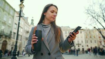 mujer vistiendo un gris Saco con un termo taza en mano caminando abajo un antiguo calle utilizando teléfono inteligente a puesta de sol. comunicación, social redes, en línea compras concepto. video