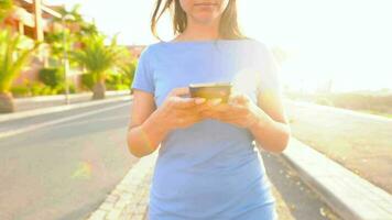 kvinna i en blå klänning använder sig av smartphone medan promenader på en handflatan gata på solnedgång video