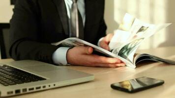 joven empresario leyendo revista por escritorio a oficina, empresario revisando y leyendo revista a gastar hora a trabajar, selectivo atención video