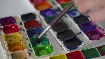 Brush takes different colors of watercolor paints from a palette and mixes them close-up video