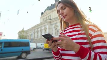 Young woman wearing a red striped sweater walking down an old street using smartphone at sunset. Communication, social networks, online shopping concept. video