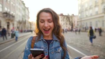 jovem mulher vestindo jeans Jaqueta caminhando baixa a velho rua usando Smartphone às pôr do sol. comunicação, social redes, conectados compras conceito. lento movimento video