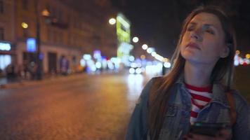 Woman is very emotionally talking with her friend on a smartphone, sees him and waves him joyfully on the street of the evening city. Blurred lights of cars and the night city on the background. video