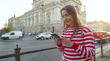 jovem mulher vestindo uma vermelho listrado suéter caminhando baixa a velho rua usando Smartphone às pôr do sol. comunicação, social redes, conectados compras conceito. video