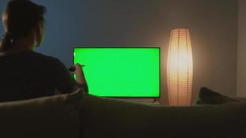 femme est séance sur le canapé, en train de regarder la télé avec une vert filtrer, commutation canaux avec une éloigné contrôle. retour voir. chrominance clé. à l'intérieur video