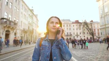 joven mujer vistiendo mezclilla chaqueta caminando abajo un antiguo calle utilizando teléfono inteligente a puesta de sol. comunicación, social redes, en línea compras concepto. lento movimiento video