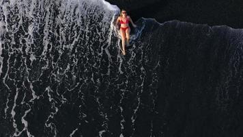 topo Visão do uma menina dentro uma vermelho roupa de banho deitado em uma Preto de praia em a surfar linha. costa do a ilha do tenerife, canário video