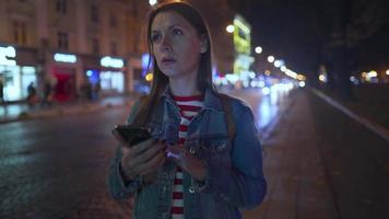 mujer es muy emocionalmente hablando con su amigo en un teléfono inteligente, ve él y olas él alegremente en el calle de el noche ciudad. borroso luces de carros y el noche ciudad en el antecedentes. video