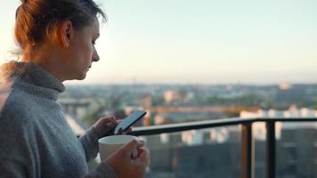 mujer empieza su día con un taza de té o café y comprobación correos electrónicos en su teléfono inteligente en el balcón a amanecer, lento movimiento. moderno urbano estilo de vida video