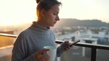 mujer empieza su día con un taza de té o café y comprobación correos electrónicos en su teléfono inteligente en el balcón a amanecer, lento movimiento. moderno urbano estilo de vida video
