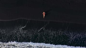 Top view of a girl in a red swimsuit lying on a black beach on the surf line. Coast of the island of Tenerife, Canary video