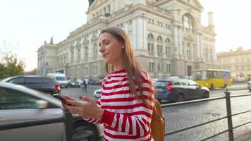 Jeune femme portant une rouge rayé chandail en marchant vers le bas un vieux rue en utilisant téléphone intelligent à le coucher du soleil. communication, social les réseaux, en ligne achats concept. video