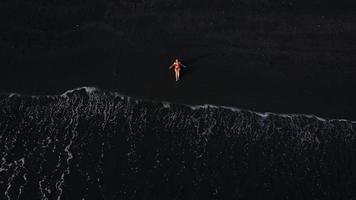 oben Aussicht von ein Mädchen im ein rot Badeanzug Lügen auf ein schwarz Strand auf das Surfen Linie. Küste von das Insel von Teneriffa, Kanarienvogel video