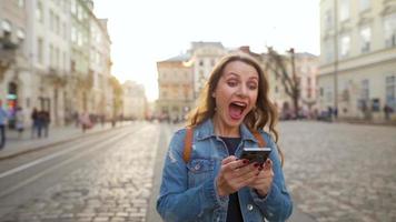 jovem mulher vestindo jeans Jaqueta caminhando baixa a velho rua usando Smartphone às pôr do sol. comunicação, social redes, conectados compras conceito. lento movimento video