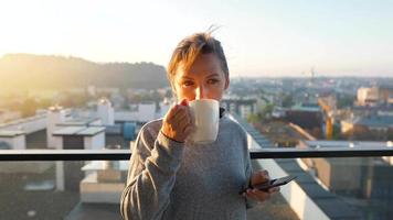 Woman starts her day with a cup of tea or coffee and checking emails in her smartphone on the balcony at dawn, slow motion. Modern urban lifestyle video