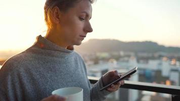 mujer empieza su día con un taza de té o café y comprobación correos electrónicos en su teléfono inteligente en el balcón a amanecer, lento movimiento. moderno urbano estilo de vida video