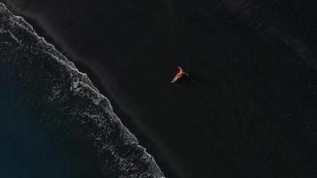 topo Visão do uma menina dentro uma vermelho roupa de banho deitado em uma Preto de praia em a surfar linha. costa do a ilha do tenerife, canário video