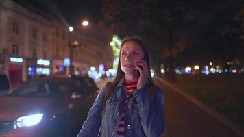 mulher é muito emocionalmente falando com dela amigo em uma Smartphone, vê ele e ondas ele alegremente em a rua do a tarde cidade. borrado luzes do carros e a noite cidade em a fundo. video