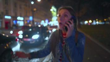 donna è molto emotivamente parlando con sua amico su un' smartphone, vede lui e onde lui con gioia su il strada di il sera città. sfocato luci di macchine e il notte città su il sfondo. video