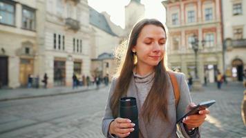 donna indossare un' grigio cappotto con un' thermos tazza nel mano a piedi giù un vecchio strada utilizzando smartphone a tramonto. comunicazione, sociale reti, in linea shopping concetto. video